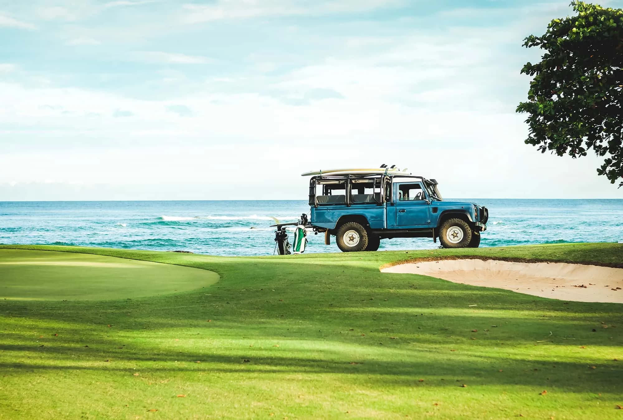 A blue Land Rover Defender with surfboards on the roof, parked above the ocean on a golf course, a golf bag leaning against it